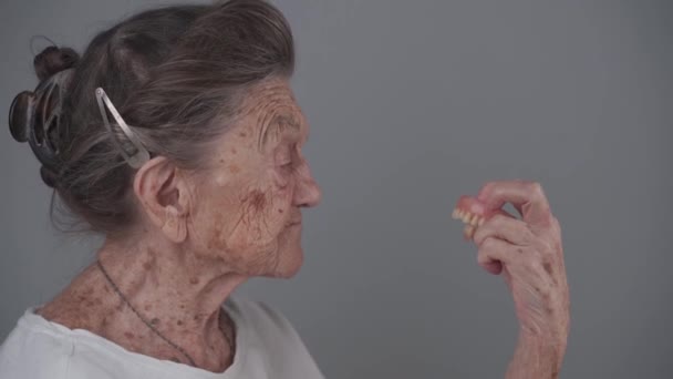 养老金领取者将假牙植入放在手上 并在工作室的灰色背景下观看 老年妇女把假牙夹在胳膊上 老年女性看着她的牙齿 牙齿植入物 — 图库视频影像