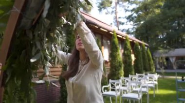 Profesyonel çiçekçi düğün kaydı için festival kemerini dekore ediyor. Kadın düğün dekoru kuruyor. Dekoratör, açık hava düğün törenini çiçekler ve yeşilliklerle süslüyor..
