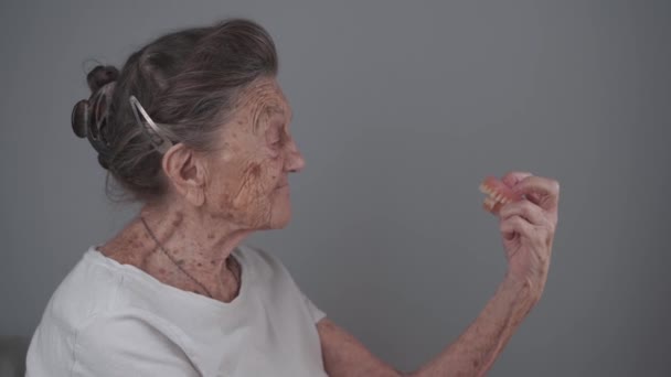 手に顎を持ち 灰色の背景を背景にスタジオを見ている老人 老婦人は入れ歯を腕につけている 年上の女性は歯を見ている 歯科インプラント — ストック動画