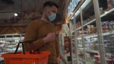 Sağlık, güvenlik alışverişi ve salgın hastalık konsepti. Maskeli bir adam marketten yiyecek alıyor. Tedbirleri alın ve engelleyin. Süpermarkette maske takan genç bir erkek el arabasıyla alışveriş yapıyor. Covid 19.
