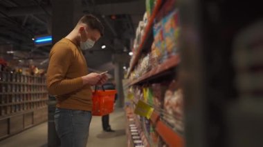 Markette koruyucu maske takan genç bir adam. Tıbbi maskeli bir erkek müşteri karantina koronavirüsü sırasında markette süt ürünlerini seçiyor. Yeni kural, maske girdisi yok.