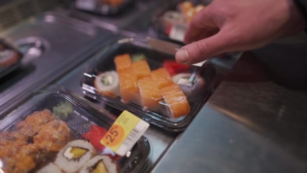 顔に保護マスクをした白人男性は 隔離とコロナウイルスの流行の間にスーパーマーケットに行くために箱の中の寿司のセットを選択します 日本の食べ物を奪う 健康食品 — ストック動画