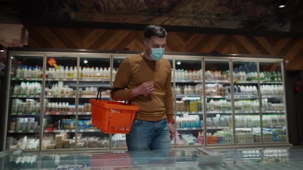 顔のマスクの若い白人男性は コロナウイルスのパンデミックの間 スーパーマーケットで乳製品を選択しています 顔文字を身に着けている食料品店での男性の買い物 手にショッピングバスケット トピック Covid — ストック動画