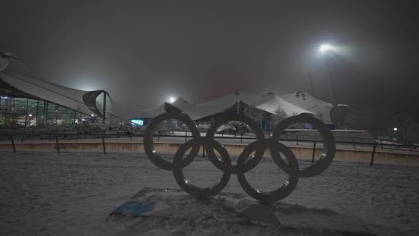 ドイツのミュンヘンのオリンピック パークで冬に雪の降る夜にオリンピックのロゴタイプ ミュンヘンの公園でオリンピアが雪で鳴っている オリンピアパーク ミュンヘン オリンピスチ リンジ — ストック動画