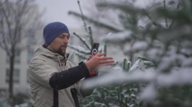 Erkek video blogcusu, sosyal ağlar için telefonuyla video çekiyor ve videodaki aboneler, kar yağışlı havalarda Noel pazarından önce sokakta bir Noel ağacı seçimi hakkında konuşuyor.. 