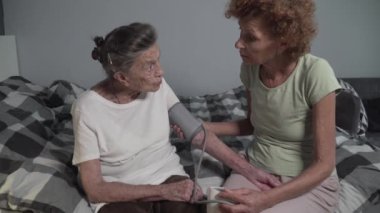 Bakıcı, evdeki yaşlı kadının tansiyon ölçümüne yardımcı oluyor. Yaşlılar sağlık kontrollerini, tansiyon monitörlerini ve sağlık hizmetlerini kullanarak yapıyor. Tıbbi kontrol. Elektronik sfinometre.