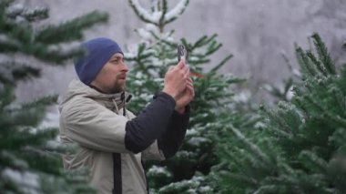 Erkek web blogcusu, sosyal medya ağları için kameralı telefon ve kışın bir Noel pazarında köknar ağacı seçmekle ilgili online yayın hizmetleri üzerinden takipçileri kullanarak video çekiyor.. 