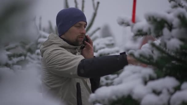 新年庆祝活动的象征 有电话购物的男人 冬天在外面的大雪中挑选 假日购物 电话交谈 询问有关选择冷杉树的建议 — 图库视频影像