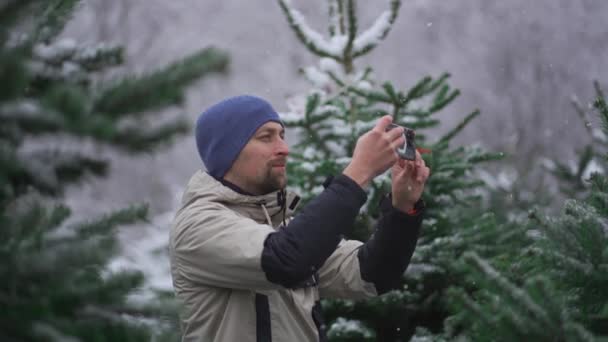 在冬季雪天的圣诞市场上 男性网络日志作者用相机手机向社交媒体网络和粉丝们拍摄有关选择冷杉树的视频 — 图库视频影像