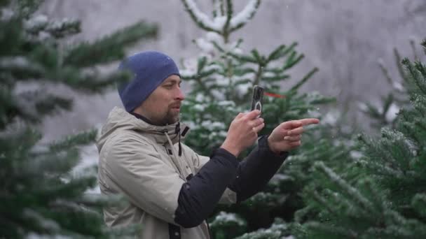 男子视频博客作者在自己的手机上为社交网络和视频订户拍摄视频 内容是在雪天的圣诞市场前 在街上选择一棵圣诞树 — 图库视频影像