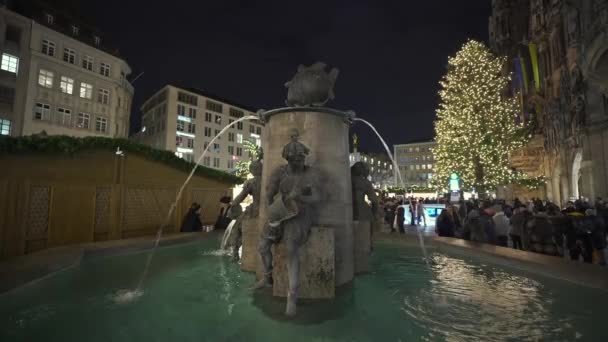 フィッシュブルネンAm Marienplatz Munchen Bayern Deutschland 2022年12月15日 クリスマスフェア中の冬の夕方には魚の噴水 背景に照らされたクリスマスツリー クリスマスマーク — ストック動画
