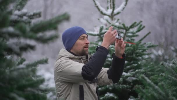 男のビデオブロガーは 雪の日にクリスマスマーケットの前に路上でクリスマスツリーの選択についてのビデオサービス上のソーシャルネットワークや加入者のための彼の電話でビデオを撮影します — ストック動画