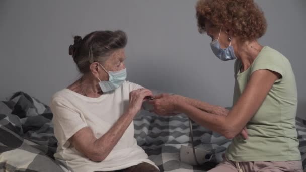 大流行時の高齢患者の血圧をチェックする医療マスクの熟女介護者 老人ホームでベッドに座って ホームケアの専門家が訪問したシニア女性 高血圧の老人 — ストック動画