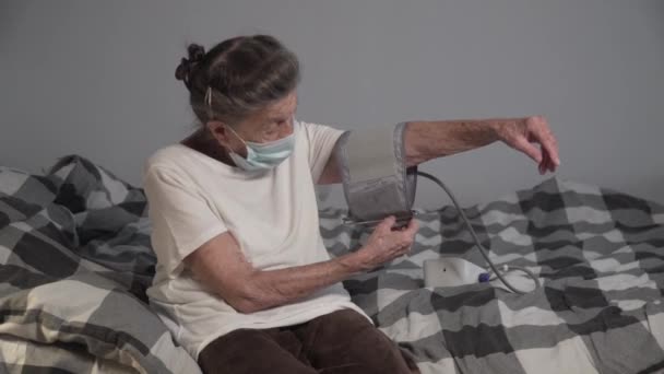 発生時に高齢女性の血圧を測定顔マスクを身に着けている熟女コロナウイルスやインフルエンザ 介護者は患者のために電子速度計を使用しています 血圧計 — ストック動画