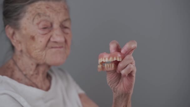 老婦人は取り外し可能な入れ歯を手に入れ カメラに映っています 口腔ケア 偽の歯と古い時代の概念 完全取り外し可能な義肢で手の高齢者の女性とともに白髪でスタジオグレーの背景 — ストック動画