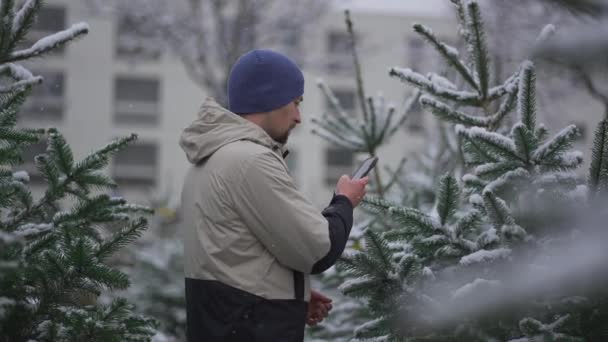ソーシャルメディアプラットフォームと冬の雪の天気でストリートネイティブ市場でのクリスマスツリーの彼の選択のオンラインストリーミングサービスのフォロワーのための彼のスマートフォン上のビデオVloggerフィルム映像 — ストック動画