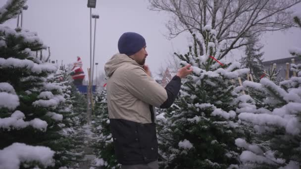 Mężczyzna Wybiera Choinkę Targu Śniegu Rozmawia Przez Telefon Żoną Doradza — Wideo stockowe