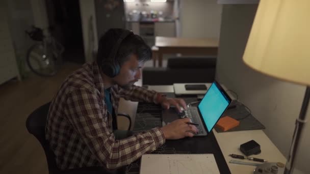 一名男生晚上带着耳机在笔记本电脑上学习 并用钢笔在笔记本上记笔记 男人深夜在电脑前工作 房间里有耳机 — 图库视频影像