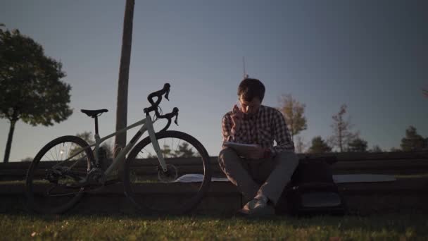 サイクリング後 大学キャンパスの公園で宿題をメモする大学生 自転車利用者はノートブックの庭でノートを取ります 偉大な屋外で勉強します 健康的なアクティブライフスタイル — ストック動画