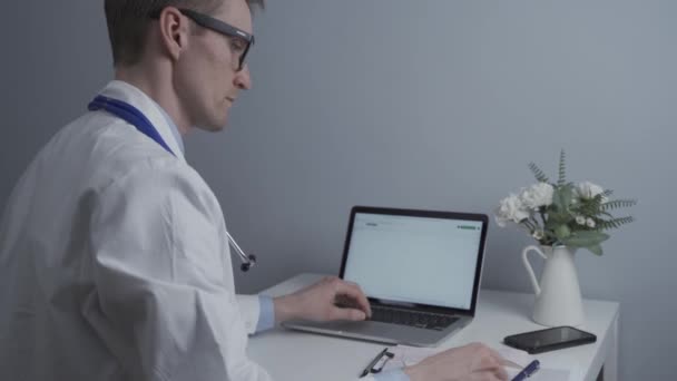 医療現場でコンピュータで働く男性医師 患者検査 ノートブックでのメモ作成 メガネとホワイトコートのクリニックでドキュメントを扱う一般医師 — ストック動画
