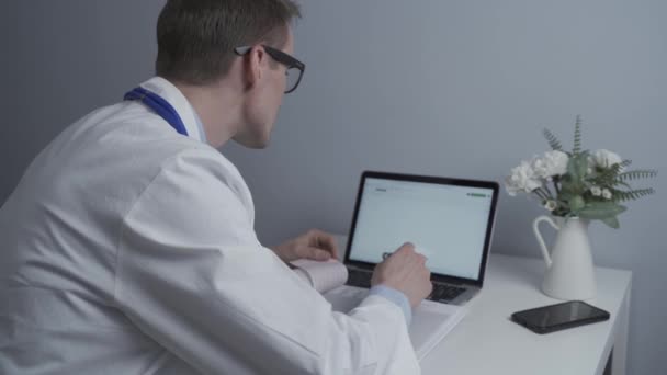 医療現場でコンピュータで働く男性医師 患者検査 ノートブックでのメモ作成 メガネとホワイトコートのクリニックでドキュメントを扱う一般医師 — ストック動画