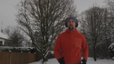 Man jogging on snowy path through park in city listening to music in earphones in winter in Germany. Athletic man working out wearing headphones in freezing cold weather. Running in snow in winter. 