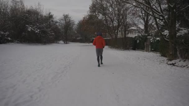 オレンジ色のスウェットシャツのバックビューで冬に雪で走る男性のジョギング 日常のアウトドアアクティブライフスタイル スポーツを楽しむ 冬に走る男のトレイルのリアビュー 凍結する雪の天気で走る — ストック動画