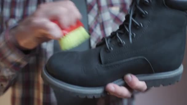 スタジオのブラシで黒いブーツを掃除する男 男性は小さなスクラブブラシでヌバックの靴をブラッシング 冬の靴のケアのテーマ 汚れた黒い秋のブーツ ワークショップの靴をきれいに洗う — ストック動画