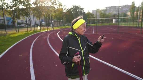 在阳光明媚的秋天里 老妇人在城市体育馆练习北欧人走路时 用的是智能手机 90多岁的老妇人在用电线杆走路的时候 会检查她在电话应用程序中走了多少步 — 图库视频影像