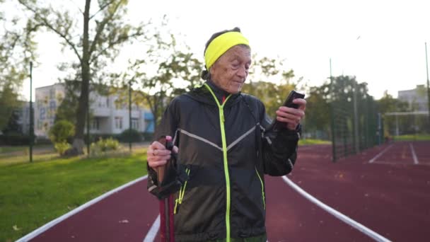 在阳光明媚的秋天里 老妇人在城市体育馆练习北欧人走路时 用的是智能手机 90多岁的老妇人在用电线杆走路的时候 会检查她在电话应用程序中走了多少步 — 图库视频影像
