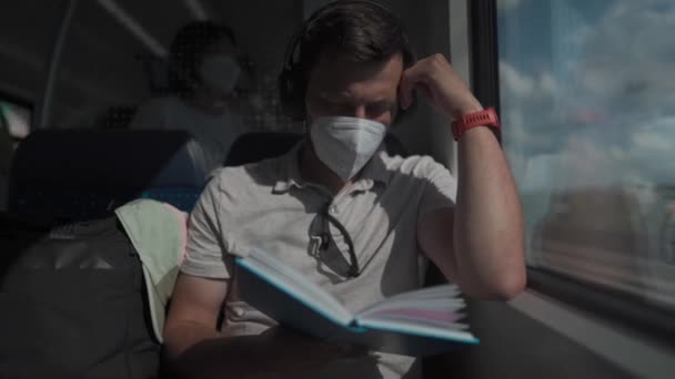 车上的白人男子戴着面具和耳机 坐在窗边看书 男人读书 在火车车厢里用耳机听音乐 穿越德国的专题铁路旅行 — 图库视频影像