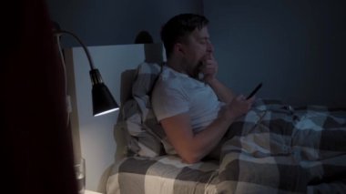 Beyaz adam uykusuzluk çekiyor, geceleri uyumuyor, internet ve akıllı telefon bağımlısı. Genç erkek gece geç saatte yastığa uzanır ve cep telefonu ekranına bakar. Yorgun gözlü adam..