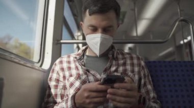 Akıllı telefon uygulaması kullanarak yüzünde KN95 FFP2 maskesi olan rahat bir adam. Tren yolu maskeli yolcu vagonda otururken ve cep telefonuyla mesajlaşırken seyahat ediyor. Toplu taşıma ile güvenli yolculuk. 
