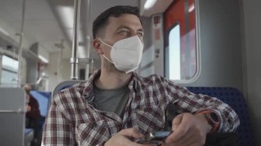 Akıllı telefon uygulaması kullanarak yüzünde KN95 FFP2 maskesi olan rahat bir adam. Tren yolu maskeli yolcu vagonda otururken ve cep telefonuyla mesajlaşırken seyahat ediyor. Toplu taşıma ile güvenli yolculuk. 