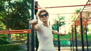 Yaşlı kadın yazın sokak fitness sahasında çalışmadan önce ısınıyor. Yaşlı bir kadın dışarıda kol ve üst vücut kemeri egzersizleri yapıyor. Emeklilik sağlıklı bir yaşam biçimi. 