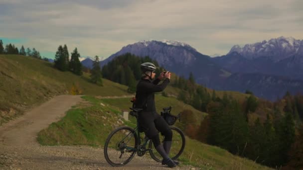 德国巴伐利亚阿尔卑斯山的男性骑自行车者在智能手机上拍照 骑自行车的人拍摄雪山覆盖的录像 骑在砾石自行车上的男性骑手欣赏风景 并在山顶上制造音响效果 — 图库视频影像