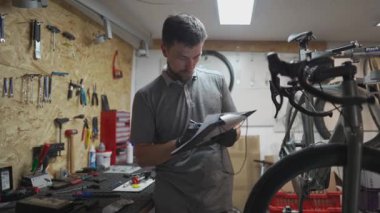 Erkek bisiklet tamircisi belgelerle çalışıyor, bisiklet atölyesinde tamamlanmış hizmetlerin kalitesini kontrol ediyor. Bisiklet teknisyeni not defterine not tutar. Mühendislik ve bisiklet bakımı. 