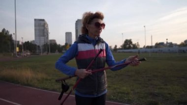 Yaşlı kadın antrenmandan önce şehir stadyumunda baston istifliyor. Olgun atletik dişi, İskandinav bastonları hazırlıyor ve ısınıyor. Kıdemli sporcu İskandinav yürüyüş direkleri için hazırlanıyor.. 