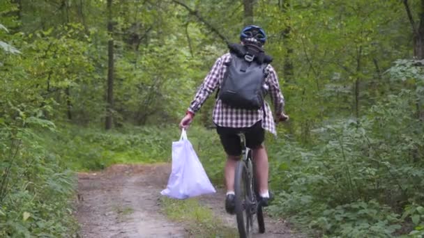 ハンドルバーにゴミ袋を持って森の中をサイクリングし 森の自転車旅行中にゴミやプラスチックを掃除します 生態系のテーマ ゴミやプラスチック廃棄物 — ストック動画