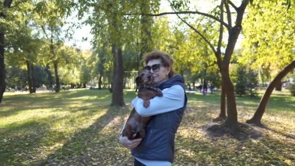 在一个阳光灿烂的秋天公园里 一个成熟的女人抱着她那只沙哑的狗 拥抱它 亲吻它 摆出姿势拍照 主题是爱宠物和户外活动 宠物女主人在户外快乐 — 图库视频影像