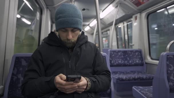 ハンブルクの地下鉄で青い座席に座り 冬の服を着たスマートフォンを使用する男性 通勤中にハンブルグの地下鉄通勤者が電話でインターネットを閲覧しています ハンバーガーUバーン — ストック動画
