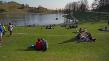 25 Mart 2022. Münih, Almanya. İnsanlar sıcak ve güneşli bahar havasında Münih 'teki Olimpik Park' ın yanındaki yeşil çimlerde dinleniyorlar. Olympiapark Münih. Olympiaberg ve Olympiasee in Olympiapark. 