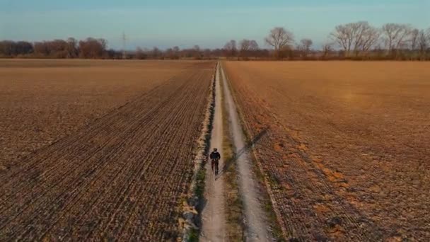 天空からの春景色の農場を介して田舎道に乗る男性のサイクリストバイク 農民が春に上陸する田舎の道を自転車で自転車でサイクリングする男 ドローンビュー — ストック動画