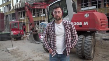 Şantiyedeki gururlu inşaatçının portresi. Kazıcılı inşaatçı yeni bir ev inşa etmeye hazır. Açık alanda ağır makinelerle duran yakışıklı bir inşaat işçisi. Mühendis