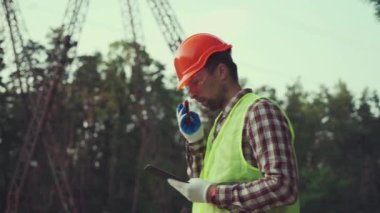 Erkek mühendis dışarıdaki elektrik hattının önünde inşaat başlığı ve yansıtıcı yelek takarak telsiz ve tablet kullanıyor. Dışarıda çalışan, radyo ve akıllı tablet kullanan bir elektrik teknisyeni.. 