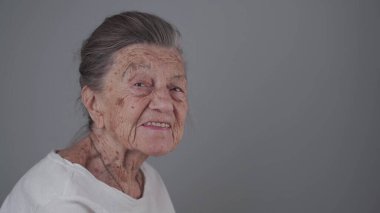 Yaşlı kadın 90 yaşında, gülümsüyor, gülüyor ve gri bir arka planda takma diş gösteriyor. Derin kırışıklıkları ve gri saçları olan yaşlı bir kadın mutlu bir şekilde stüdyo fotoğrafına bakıyor..