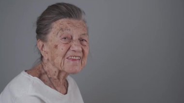 Yaşlı kadın 90 yaşında, gülümsüyor, gülüyor ve gri bir arka planda takma diş gösteriyor. Derin kırışıklıkları ve gri saçları olan yaşlı bir kadın mutlu bir şekilde stüdyo fotoğrafına bakıyor..
