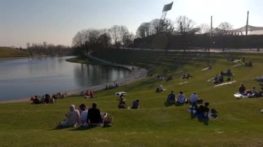 25 Mart 2022. Münih, Almanya. İnsanlar sıcak ve güneşli bahar havasında Münih 'teki Olimpik Park' ın yanındaki yeşil çimlerde dinleniyorlar. Olympiapark Münih. Olympiaberg ve Olympiasee in Olympiapark. 