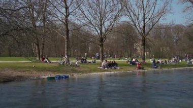 22 Mart 2022. Münih, Almanya. İnsanlar baharda güneşli ve sıcak havalarda Englischer Bahçesi 'ndeki Isar Nehri yakınlarındaki yeşil çimlerde dinleniyorlar. İnsanlar sıcak günün tadını çıkarıyorlar, Münih 'teki Isar Nehri yakınlarında güneşleniyorlar.. 