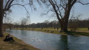 22 Mart 2022. Münih, Almanya. İnsanlar baharda güneşli ve sıcak havalarda Englischer Bahçesi 'ndeki Isar Nehri yakınlarındaki yeşil çimlerde dinleniyorlar. İnsanlar sıcak günün tadını çıkarıyorlar, Münih 'teki Isar Nehri yakınlarında güneşleniyorlar.. 